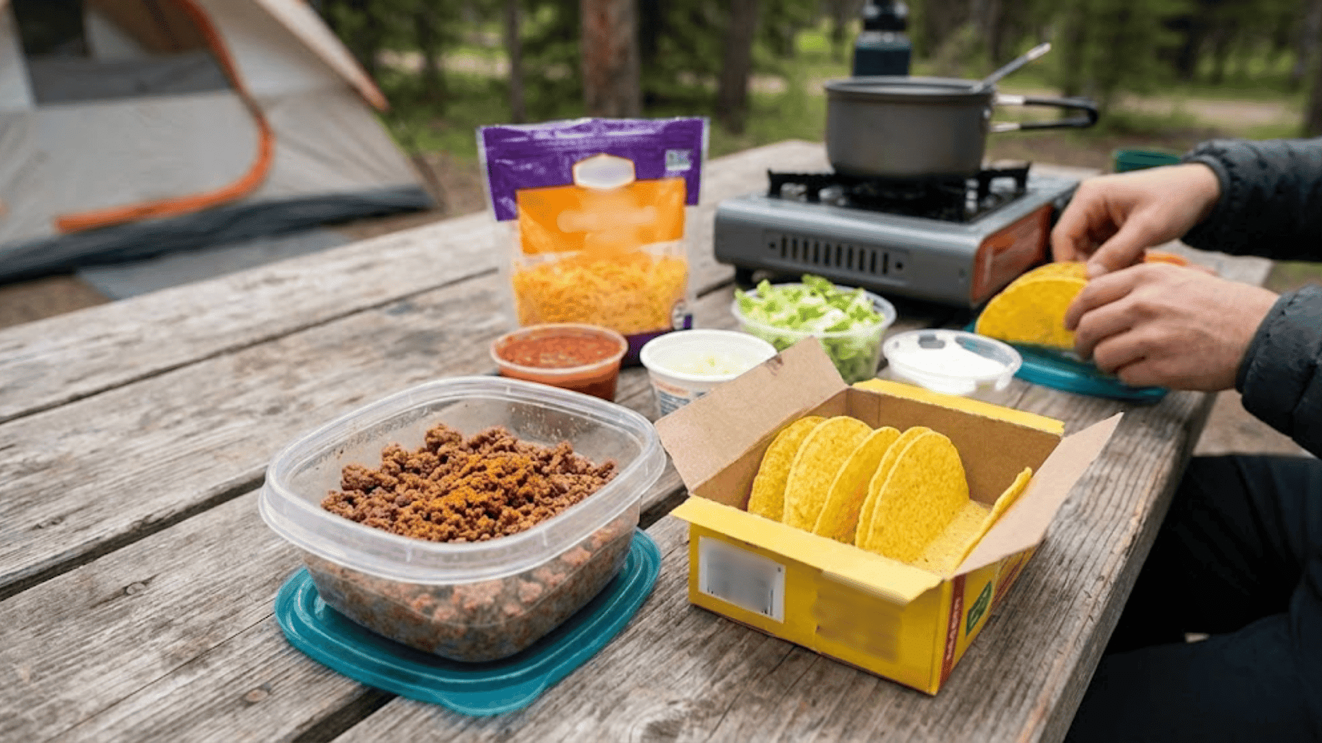 Camper preparing tacos with ground meat, cheese, and toppings on outdoor table