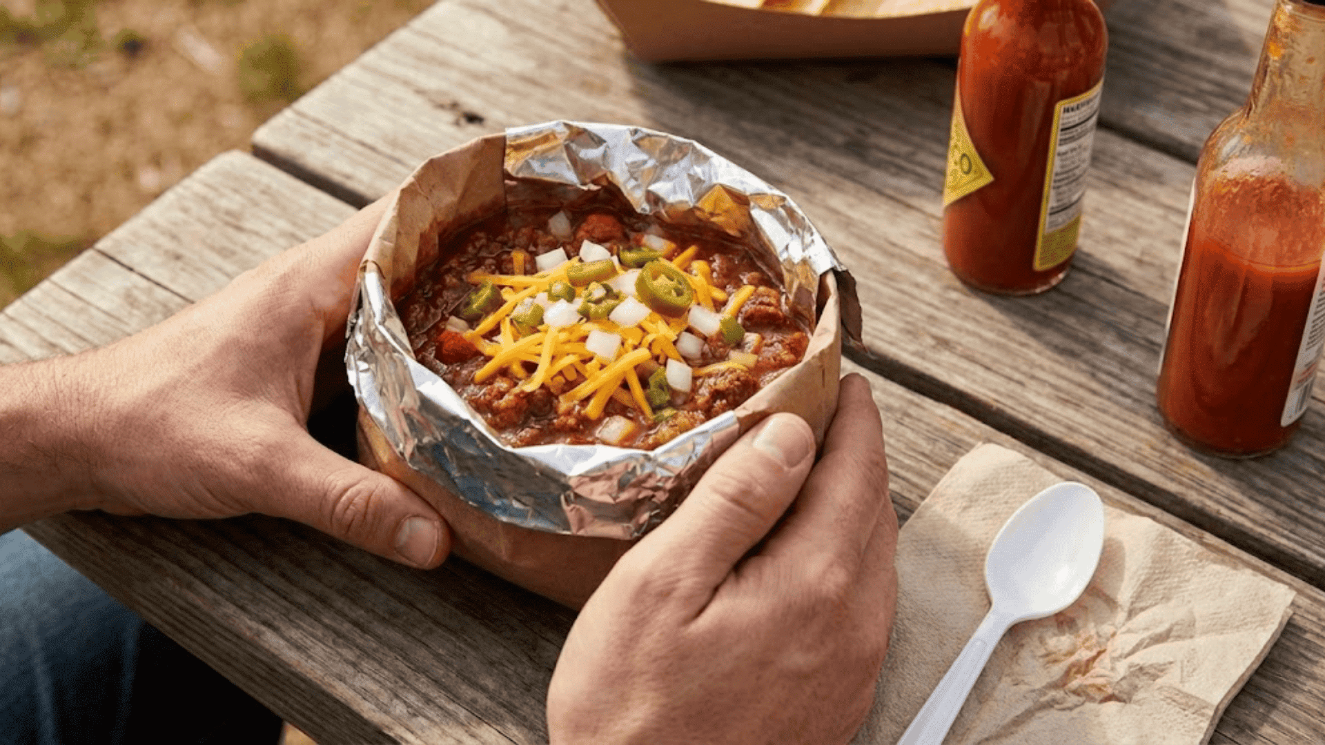 Chili served in foil-lined bowl topped with cheese, jalapeños, and onions on picnic table