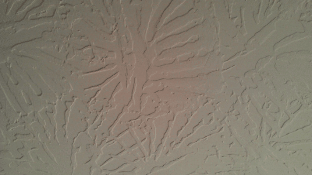 Slap brush knockdown drywall texture shows flat tan plaster peaks over a stippled base with soft side shadows on a wall