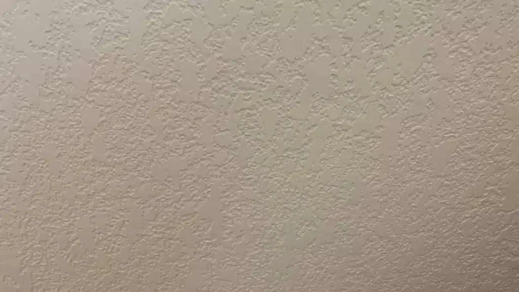 Splatter knockdown drywall texture shows flat beige plaster spots over a bumpy spray pattern with soft uniform lighting