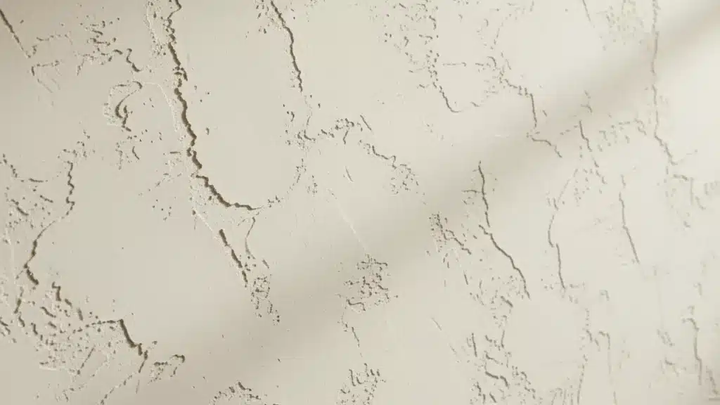 closeup of knockdown drywall texture showing flat beige peaks and mottled recessed areas with soft diagonal shadowing