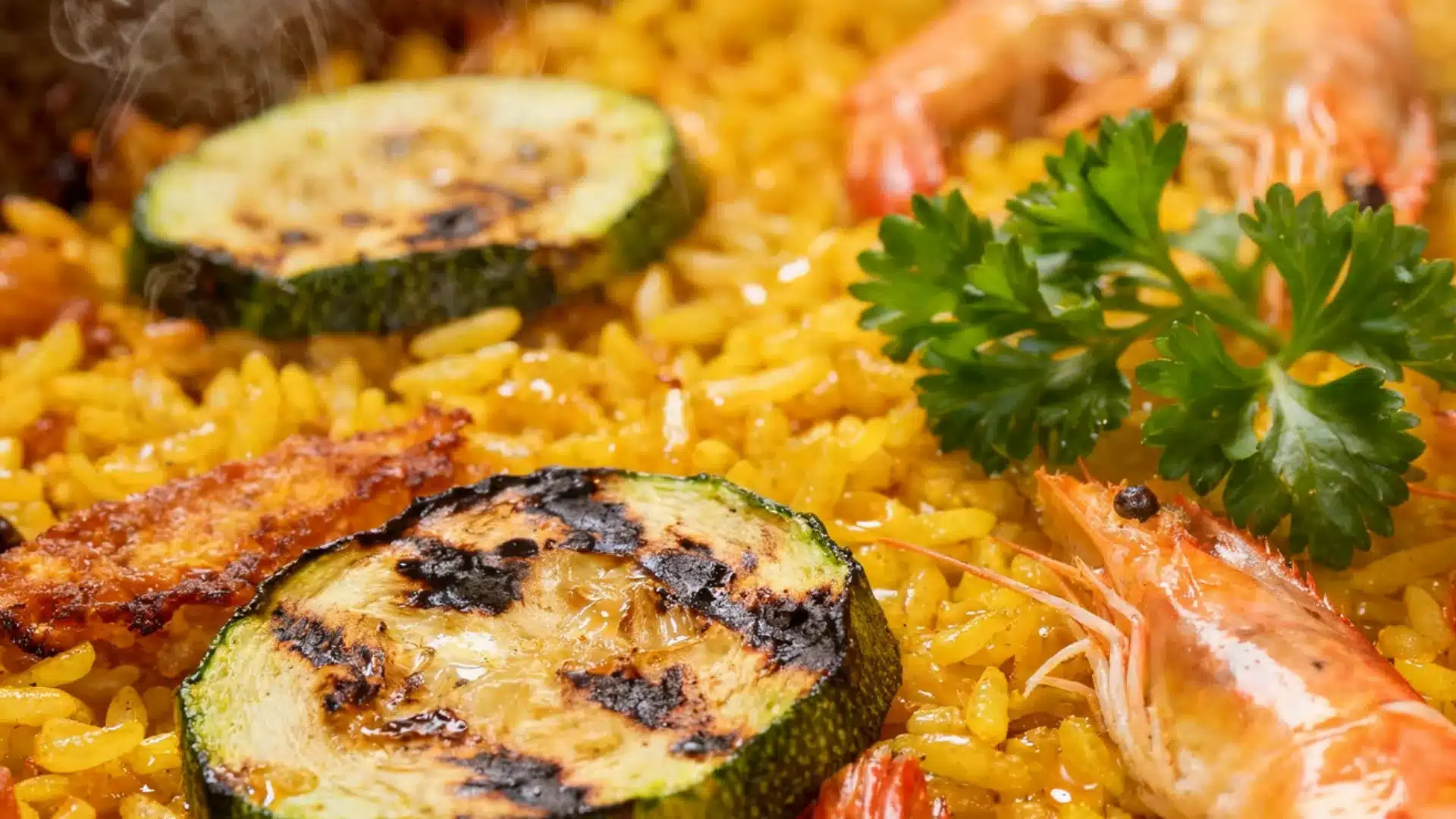 A close up of steaming yellow paella garnished with grilled zucchini slices, large shrimp, and fresh parsley