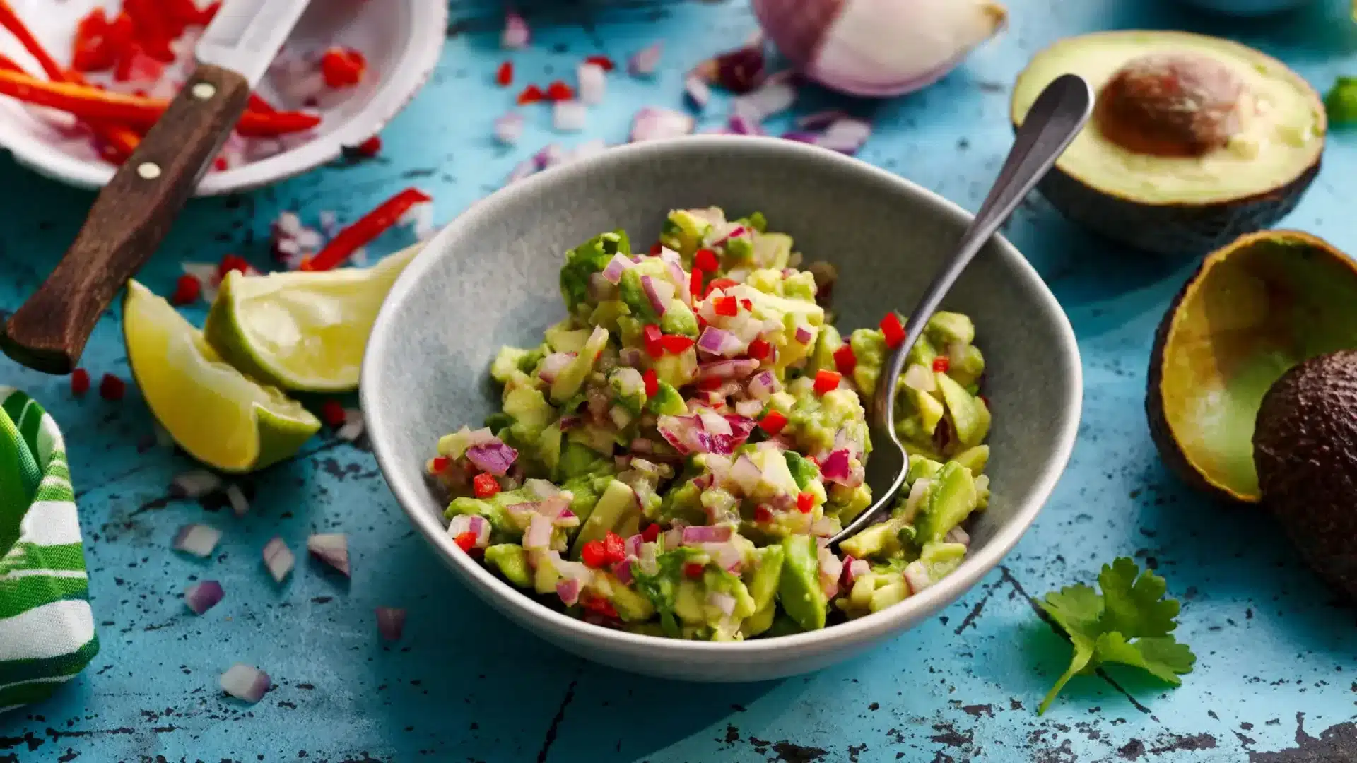 Chunky avocado salsa mixed with diced red onion and chili peppers served in a bowl on a textured blue surface with lime wedges and whole avocados nearby.