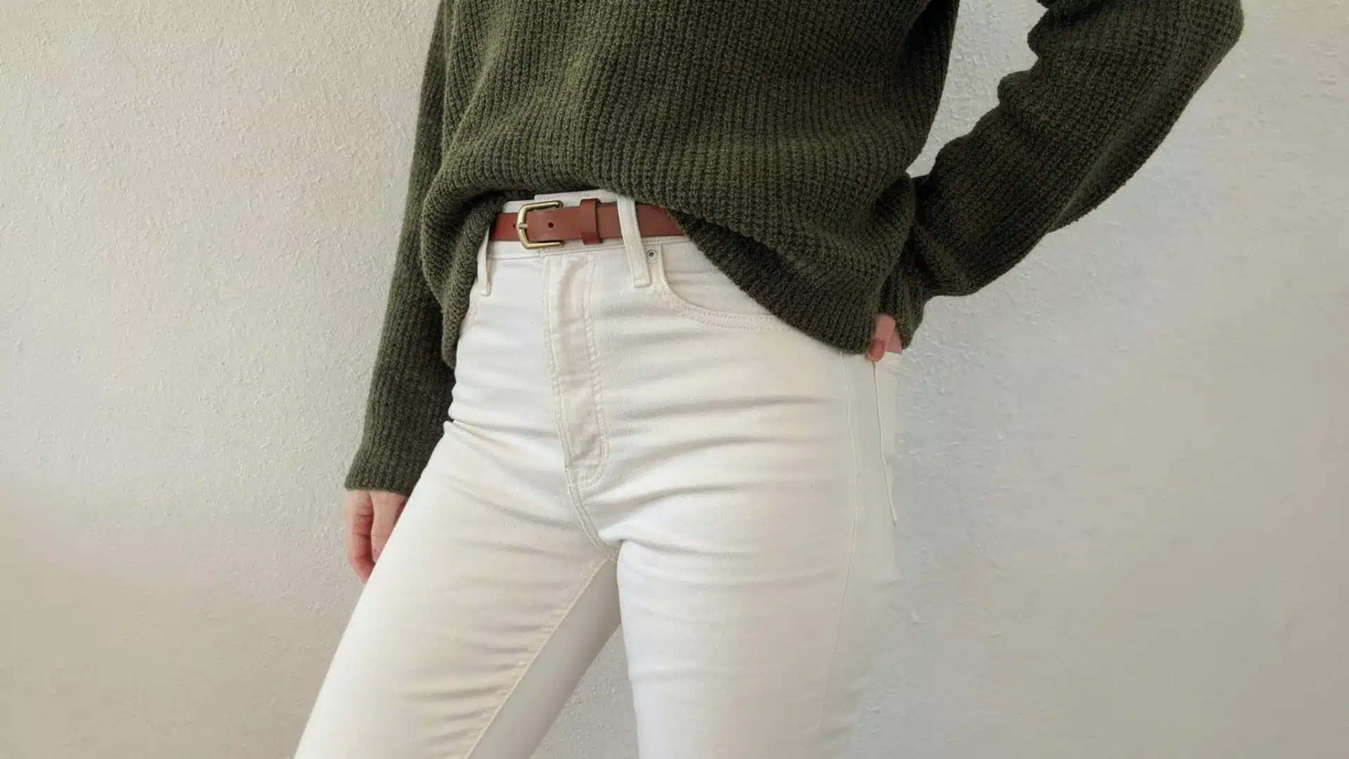 Close-up of a person wearing a dark green knit sweater tucked into high-waisted white jeans secured with a thin brown leather belt