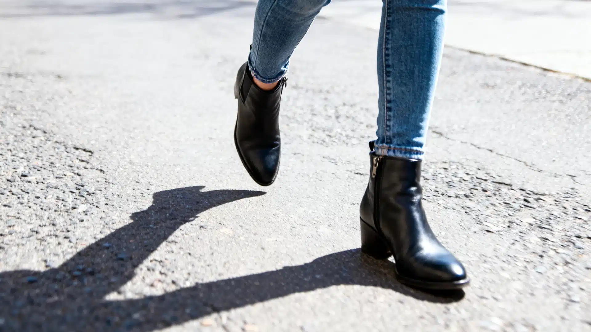 Close-up of legs wearing skinny blue jeans and black leather ankle boots walking on sunlit gray pavement with a strong shadow cast below