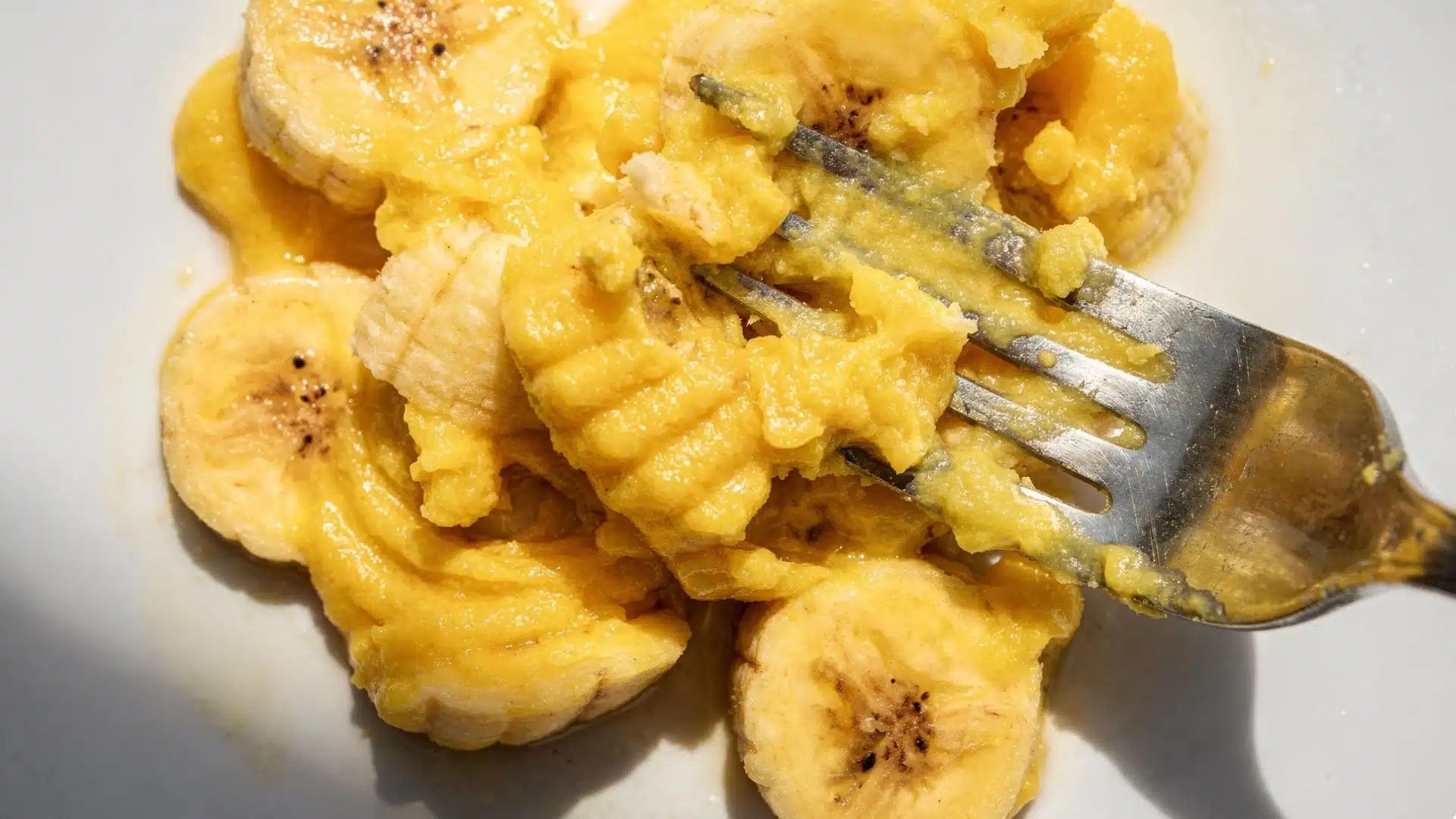Closeup overhead shot of mashed and sliced yellow bananas on a white plate being pressed down by a silver metal fork tines (1)