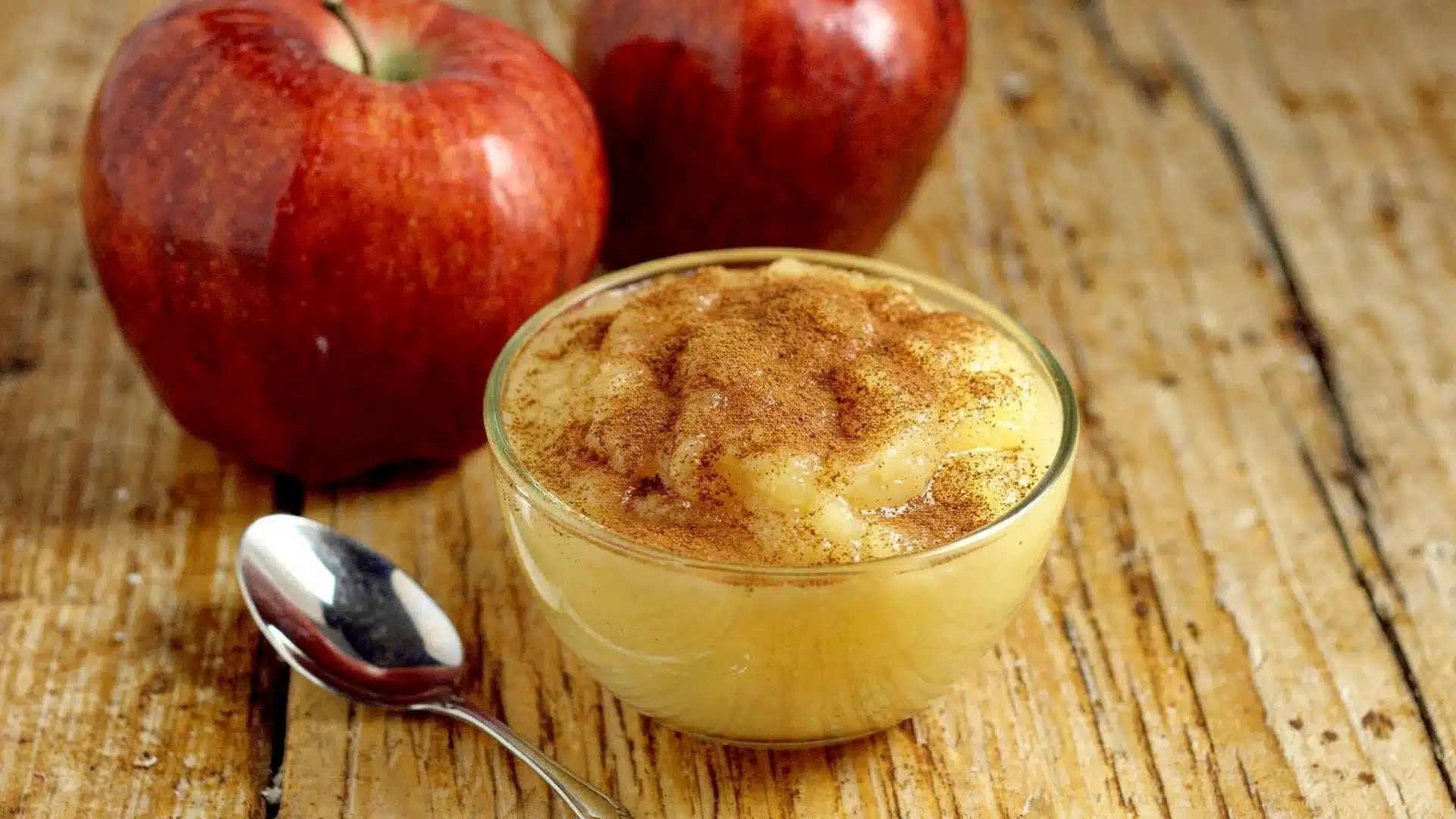 Small glass bowl of applesauce topped with cinnamon next to two red apples and a silver spoon on a rustic wooden table (1)