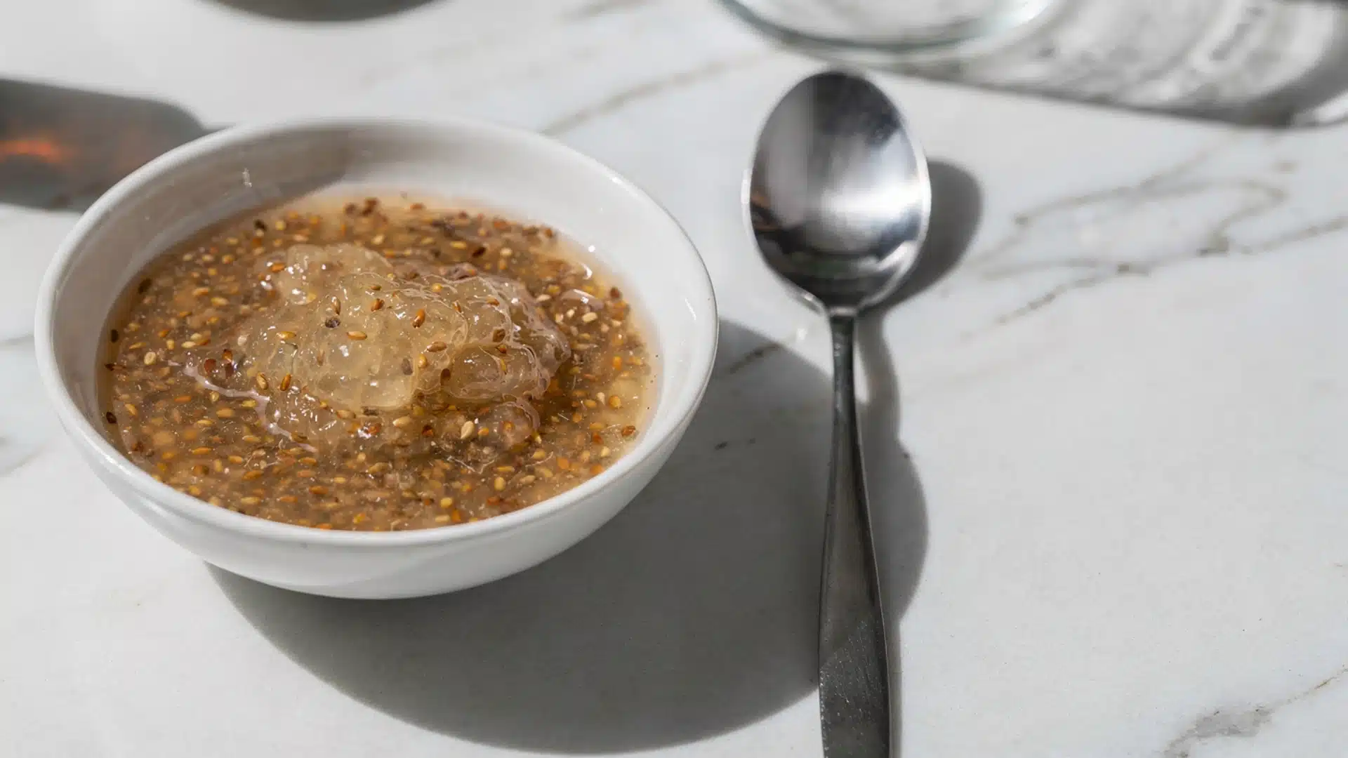 White bowl of flaxseed gel with a metal spoon amber bottle of vanilla and white powder on a marble counter in bright light (1)