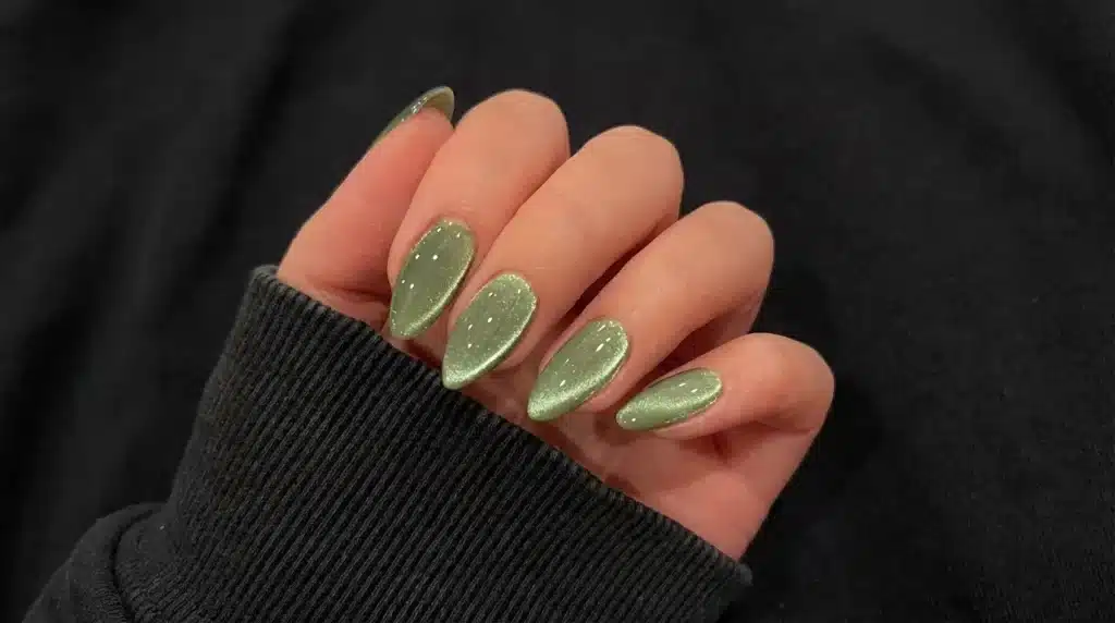 Close-up of a hand showcasing almond-shaped nails with shimmery, sage green magnetic cat-eye polish against a dark, ribbed fabric background