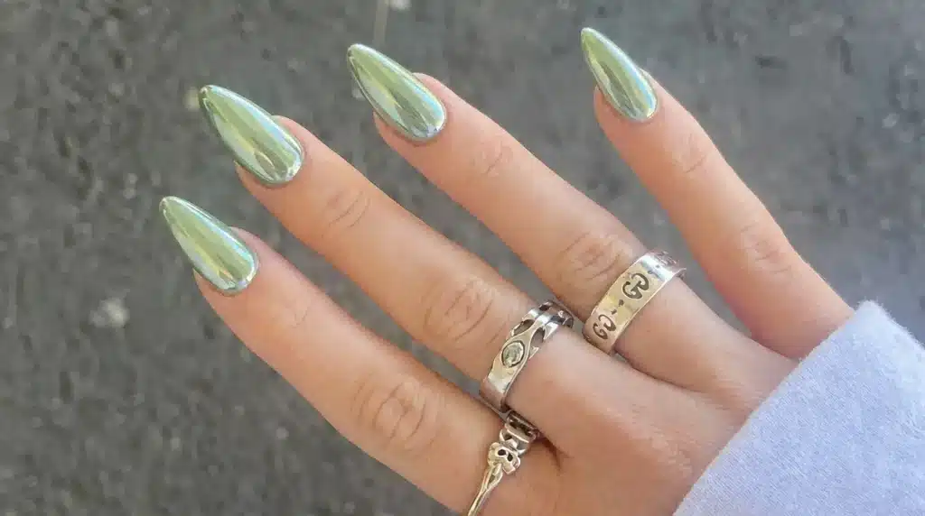 Close-up of a hand with stiletto-shaped nails painted in a reflective, chrome sage green polish, adorned with several silver-toned rings