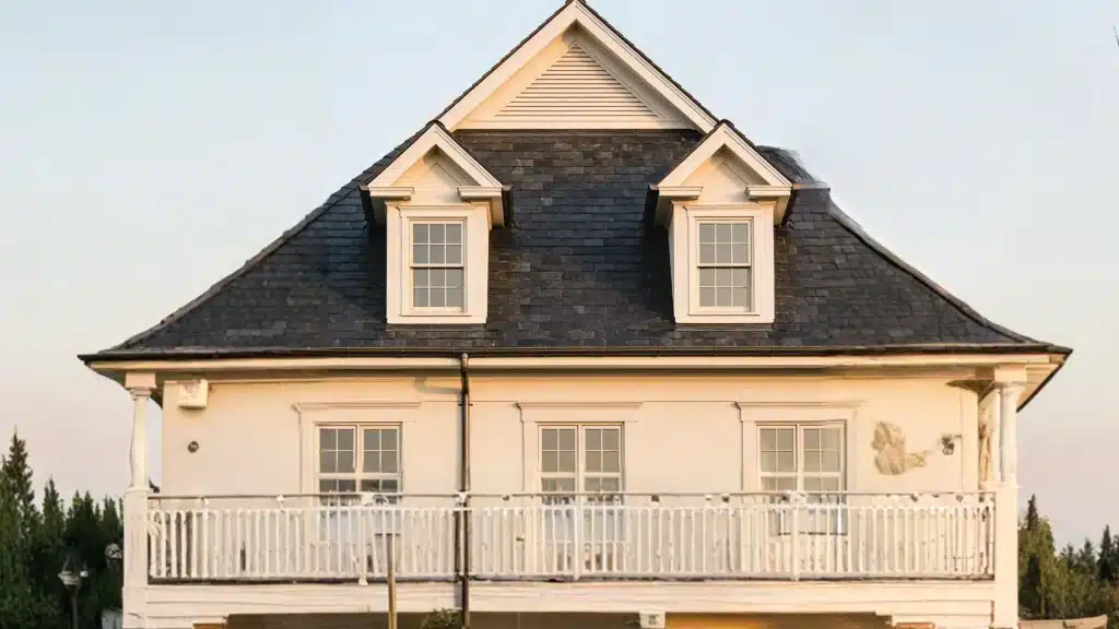 Close-up of a light-colored house facade featuring a steep dark slate roof with two white-trimmed dormer windows beneath a central gabled vent.
