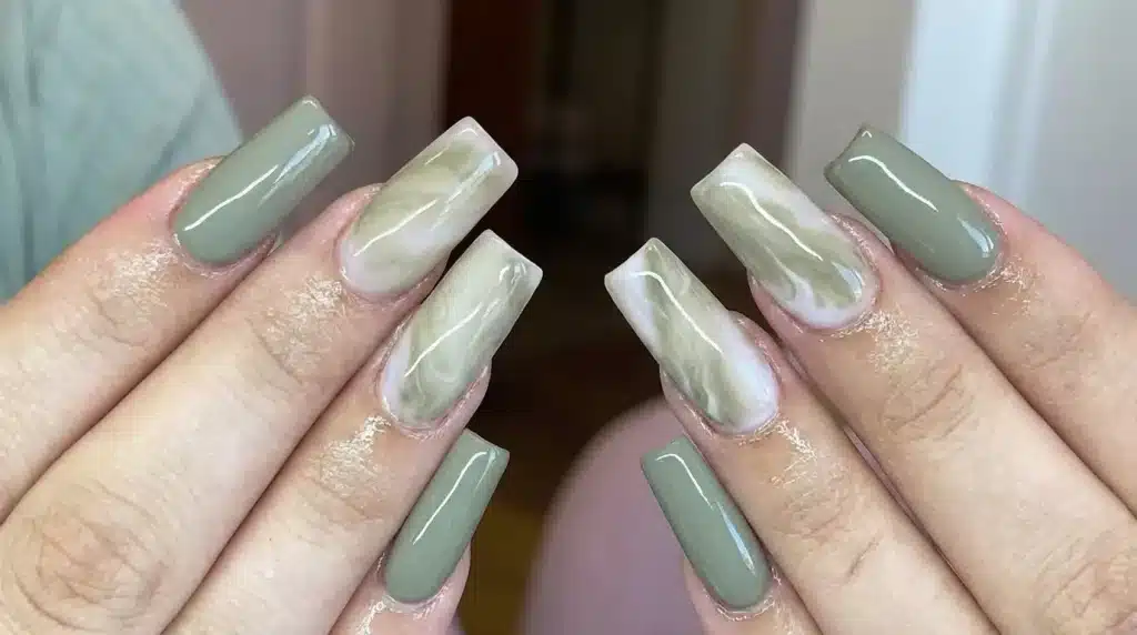 Close-up of hands showing long, coffin-shaped acrylic nails featuring alternating solid sage green polish and white and green marble design