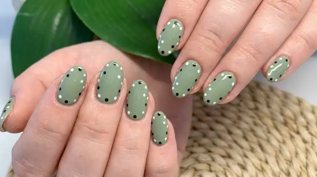 Close-up of hands showing oval-shaped nails painted sage green with scattered black and white polka dots, set against a blurred background of a green leaf and woven texture