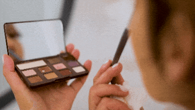 Person holding eyeshadow palette and brush, applying neutral shades while looking in the mirror, doing a makeup routine