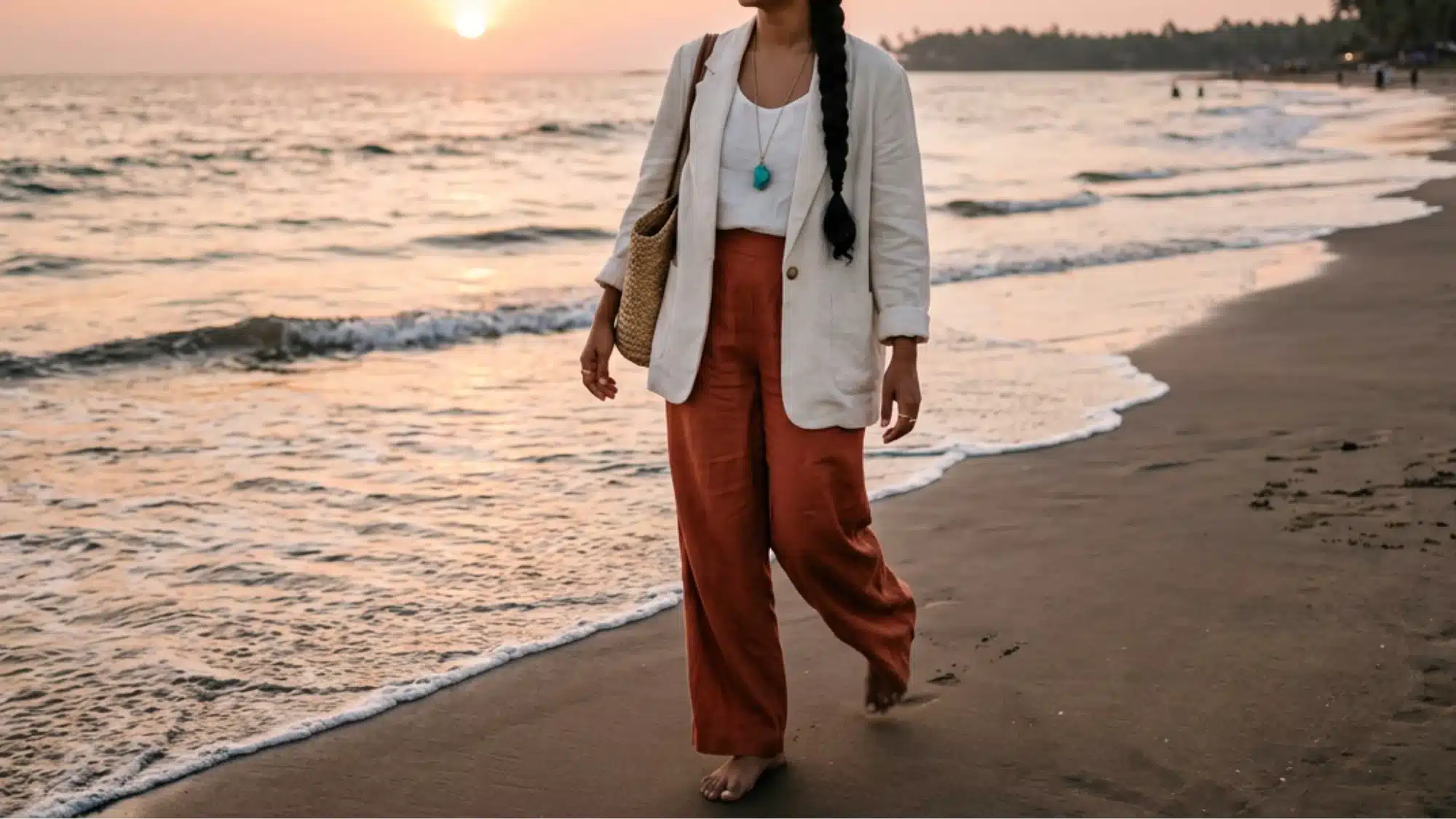 Woman in white blazer and terracotta pants walks barefoot on a sandy beach at sunset with gentle waves washing ashore