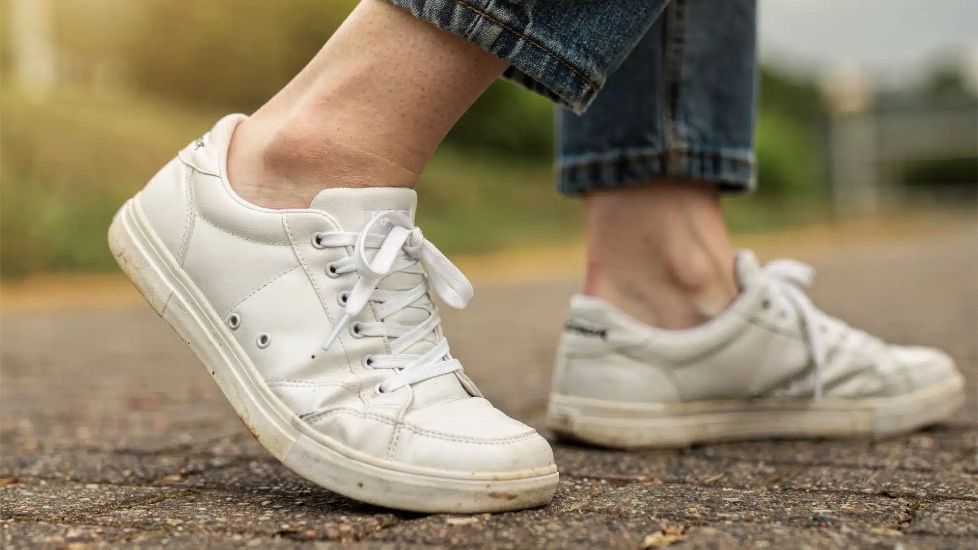 Close-up of a person wearing slightly worn white lace-up sneakers and rolled-up blue jeans while walking outdoors on a paved surface