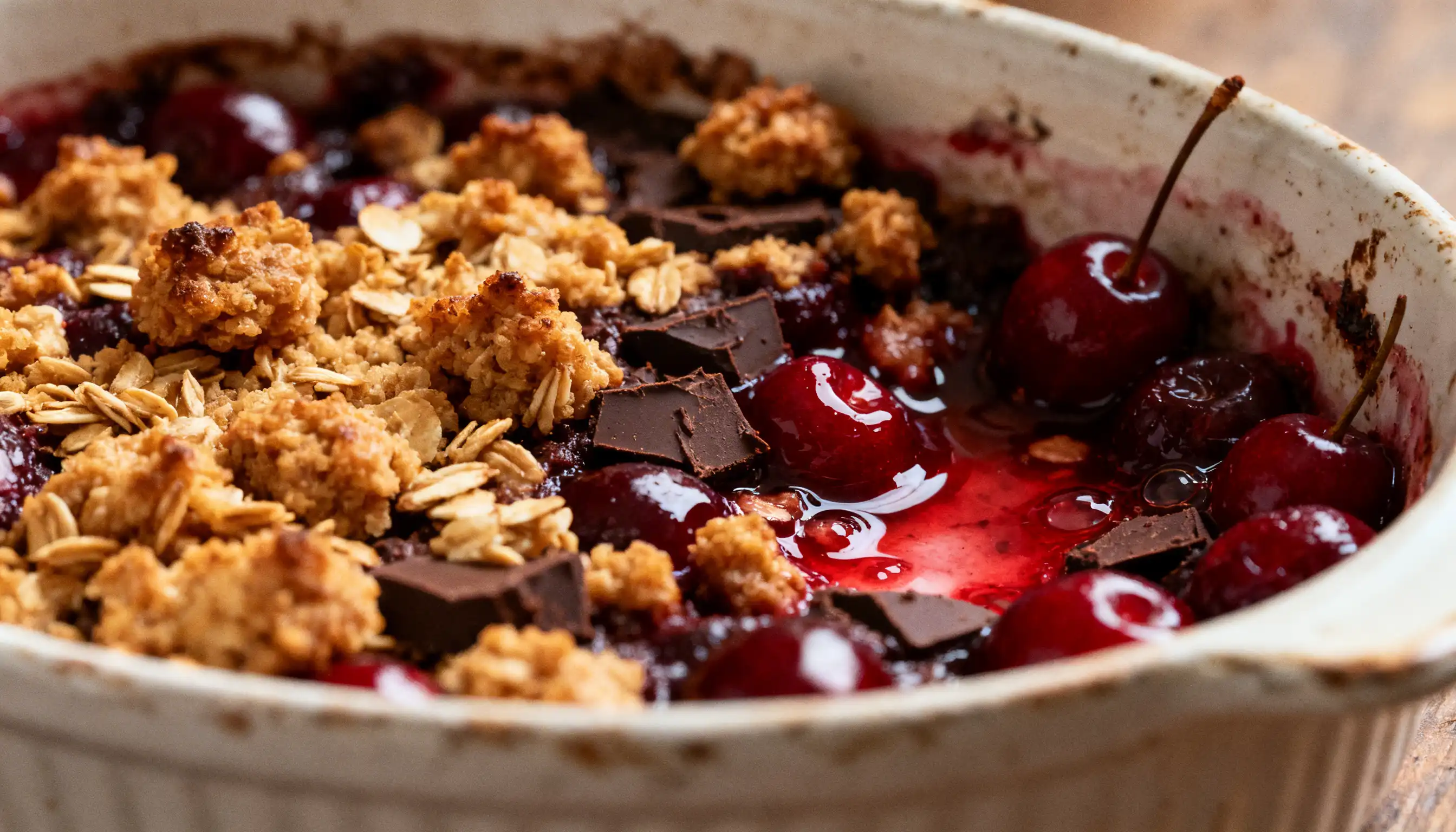 Close-up of a warm cherry and chocolate crumble topped with toasted oats, served in a rustic white baking dish.