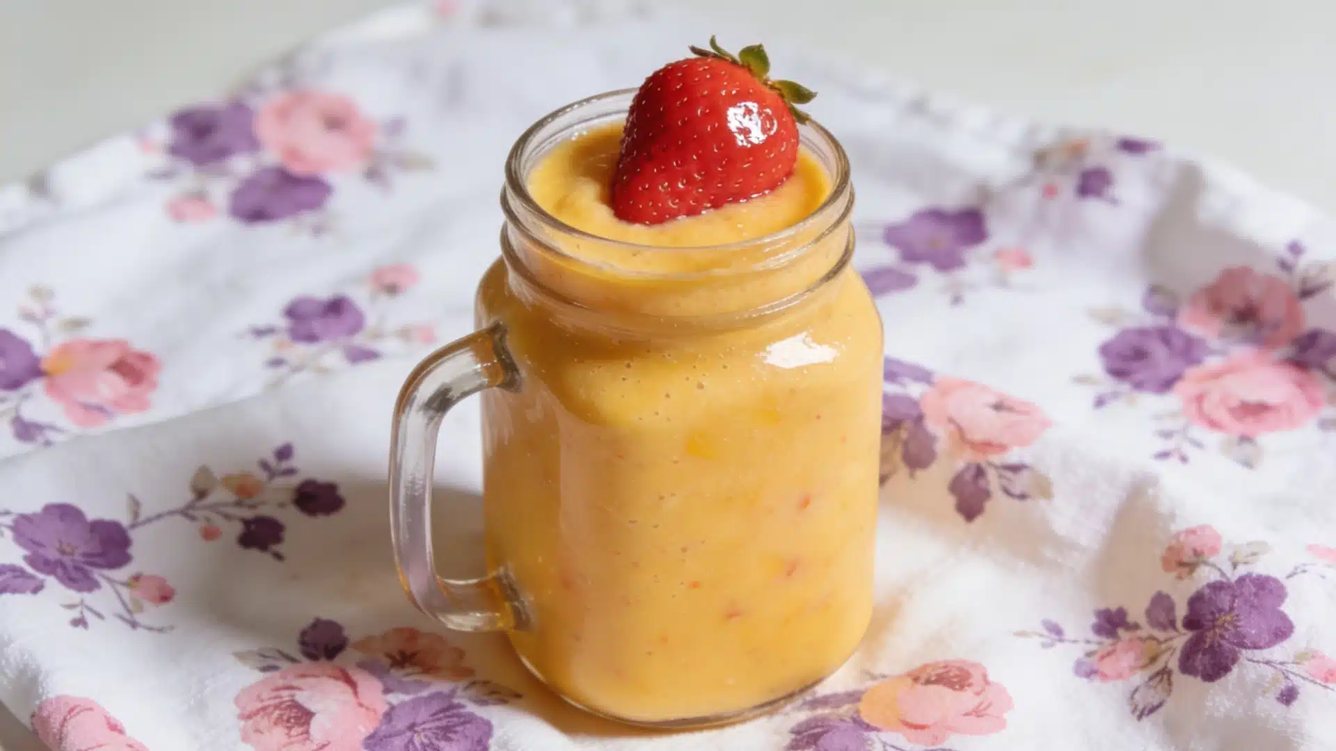A vibrant mango smoothie in a mason jar topped with a fresh strawberry, set on a floral-patterned cloth, conveying a cheerful and refreshing vibe.